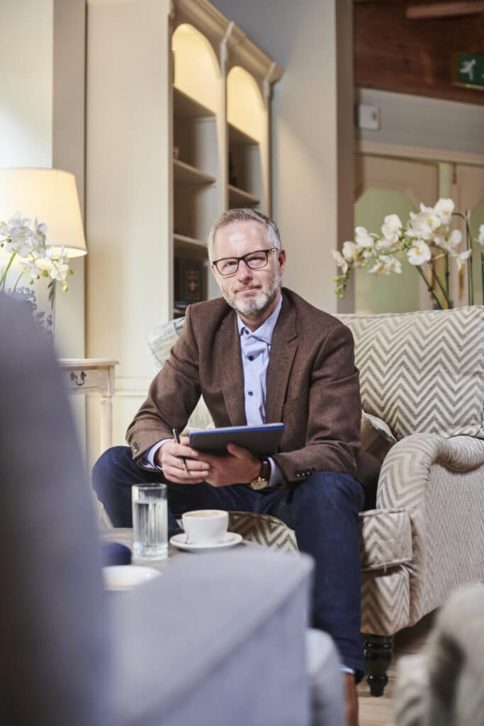 Man with glasses and brown blazer sitting on a patterned armchair holding a tablet and pen