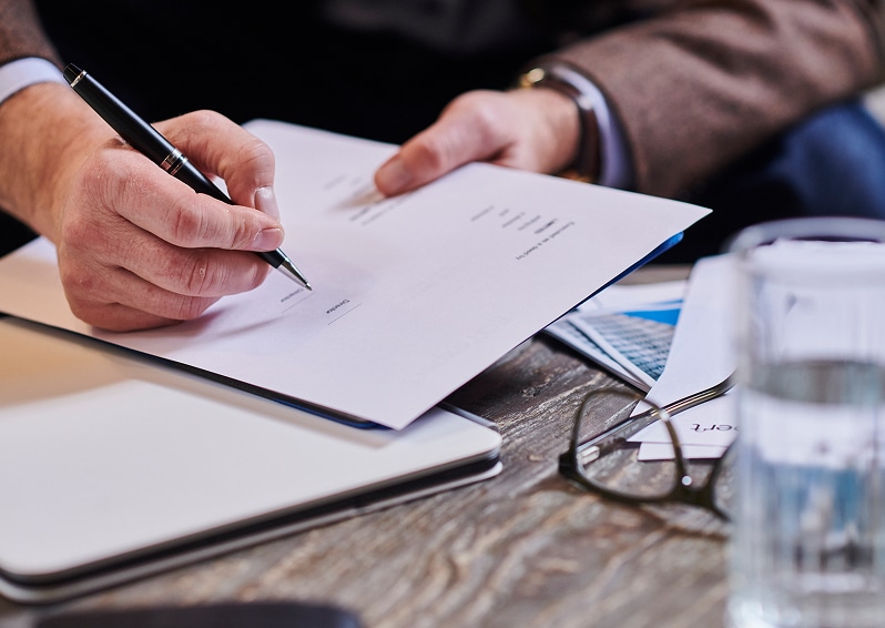 Person signing a document with a pen on a wooden table with glasses and water nearby