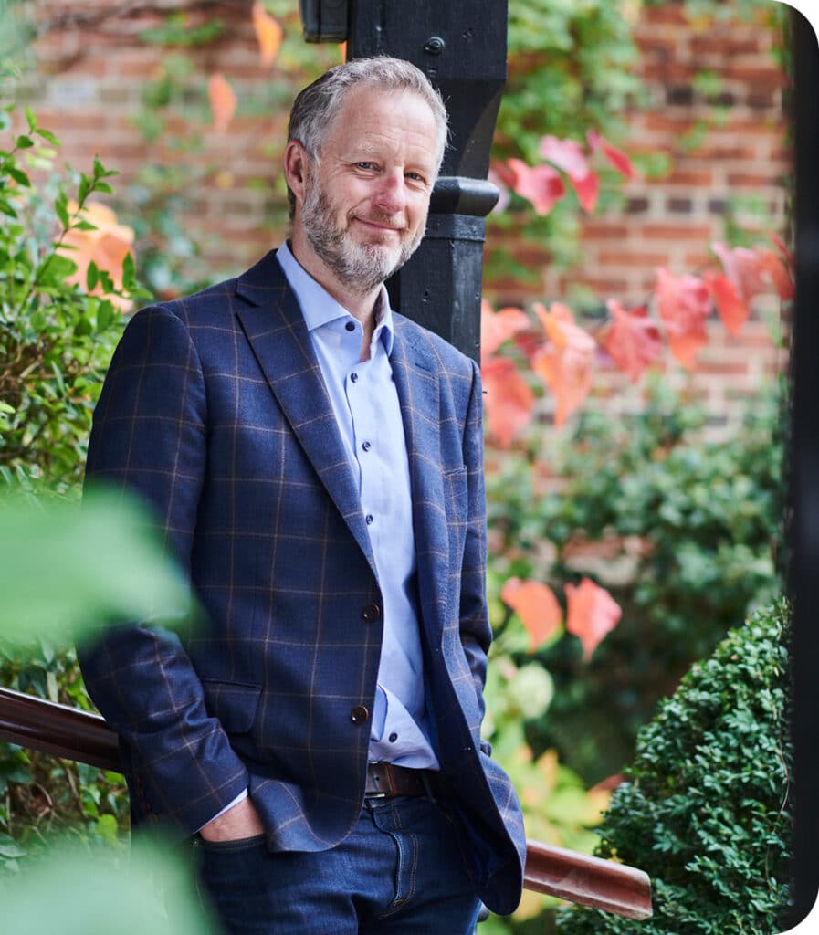 Man with grey hair and beard wearing blue checked blazer and light blue shirt standing outdoors